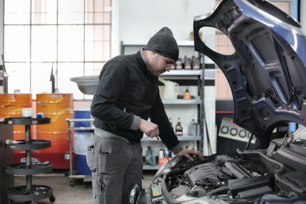A man in a blue jacket stands beside a car engine, examining its components with a focused expression.